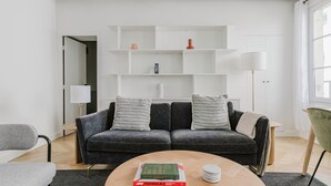 A modern living room with a grey sofa, two pillows, a coffee table with books, and white shelving units. - Blueground | Grands Blvds, washer, nr shops (Paris)
