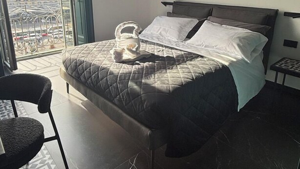 A bedroom with a bed, a chair, a small table, and a view of buildings through a large window. - Volta 70 (Naples)