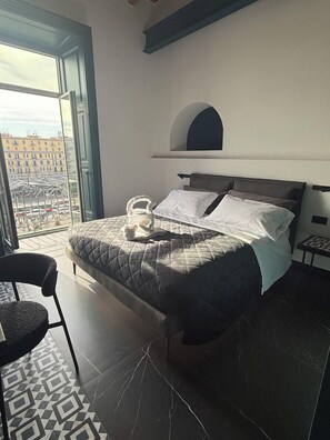 Un dormitorio con cama, una silla, una mesita y vistas a edificios a través de un ventanal grande.