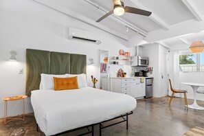 A modern bedroom with a bed, a ceiling fan, an air conditioning unit, and a kitchenette in the background.