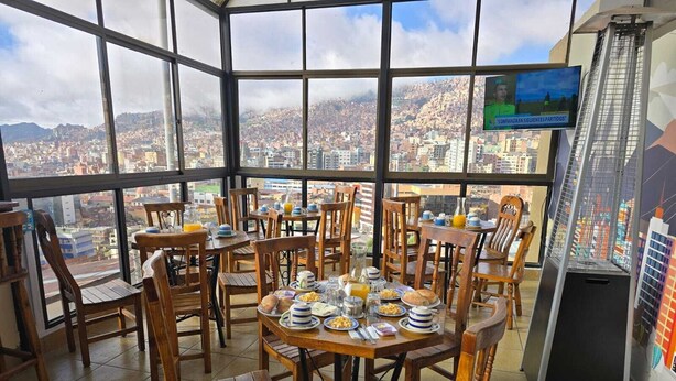 Un comedor con varias mesas y sillas, alimentos para el desayuno y vistas de la ciudad a través de amplios ventanales.