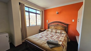 Un dormitorio con cama, ventana con cortinas, mesita de noche y lámpara de pared.