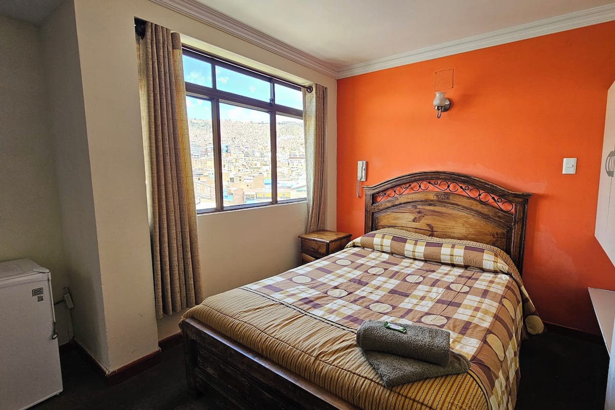 A bedroom with a bed, a window with curtains, a nightstand, and a wall-mounted light fixture.