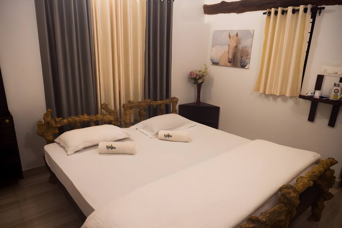 A double bed with a wooden headboard, white linens, and two towels with a logo. A small bedside table with a vase of flowers is next to the bed.