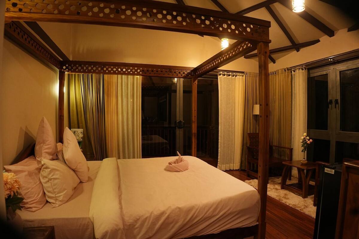 A four-poster bed with a canopy, a small table with a vase of flowers, and a view of the outside through a glass door.
