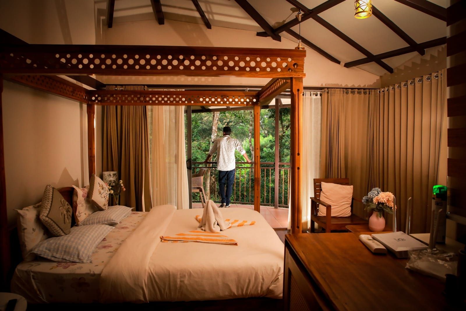 A bedroom with a bed, curtains, a wooden chair, a table with a vase, and a balcony with a person.