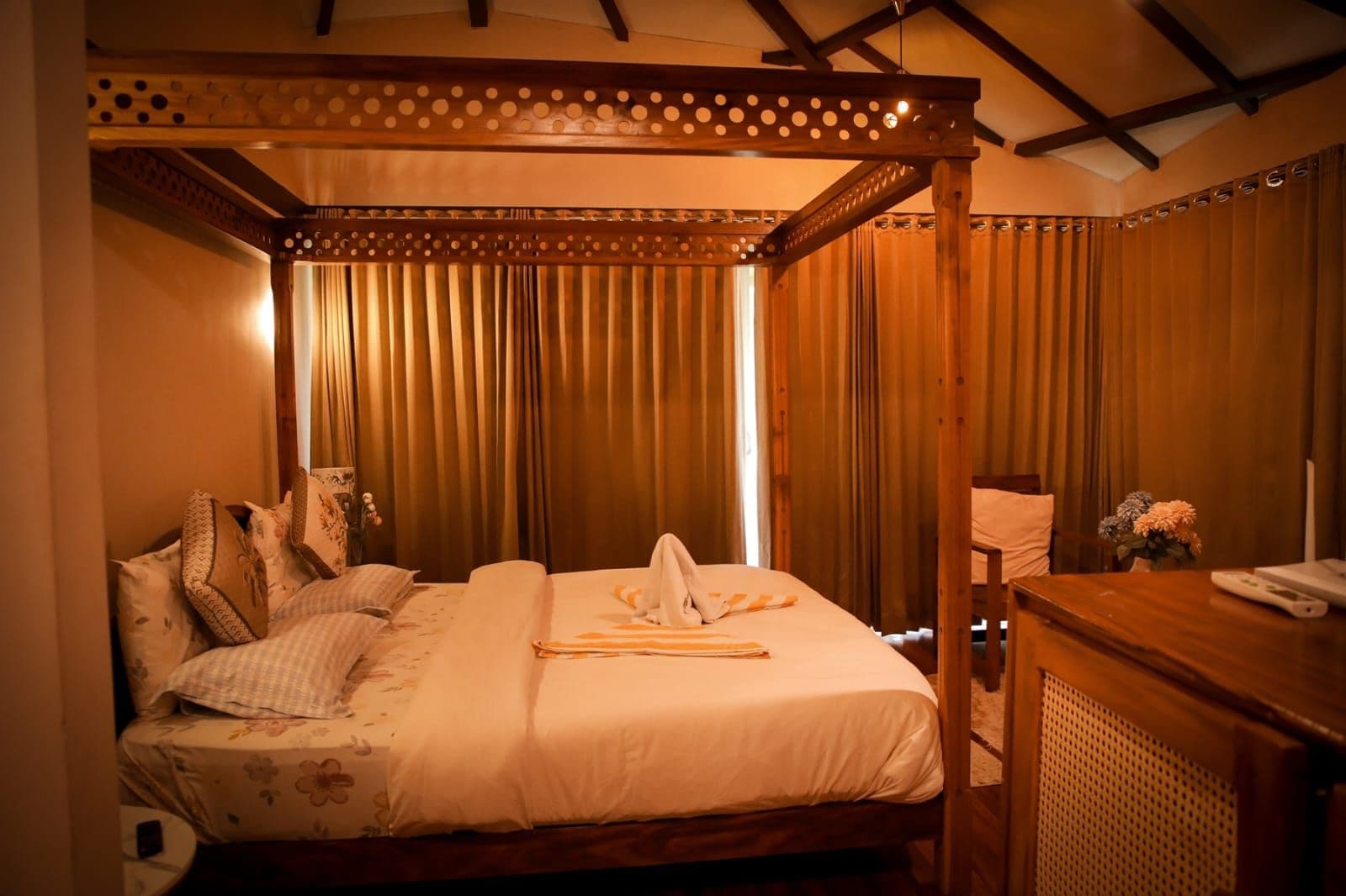 Una cama con dosel, una mesita de noche con un teléfono y un florero con flores.
