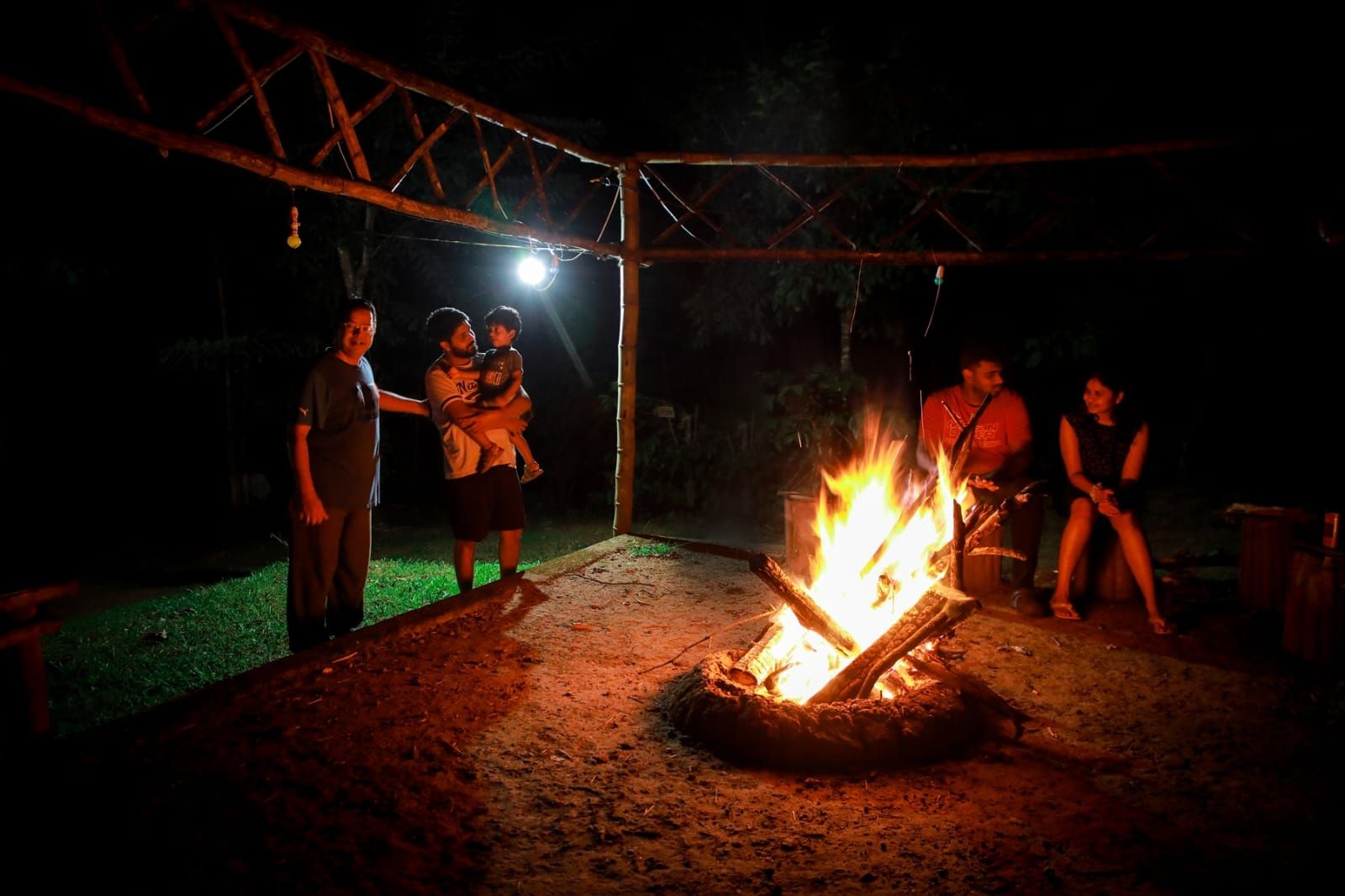 An outdoor gathering around a campfire with a metal structure overhead.