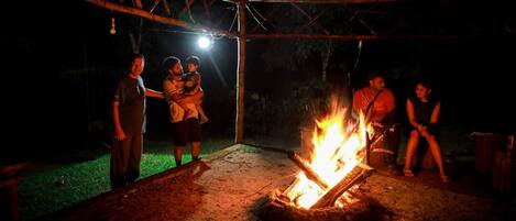 An outdoor gathering around a campfire with a metal structure overhead.