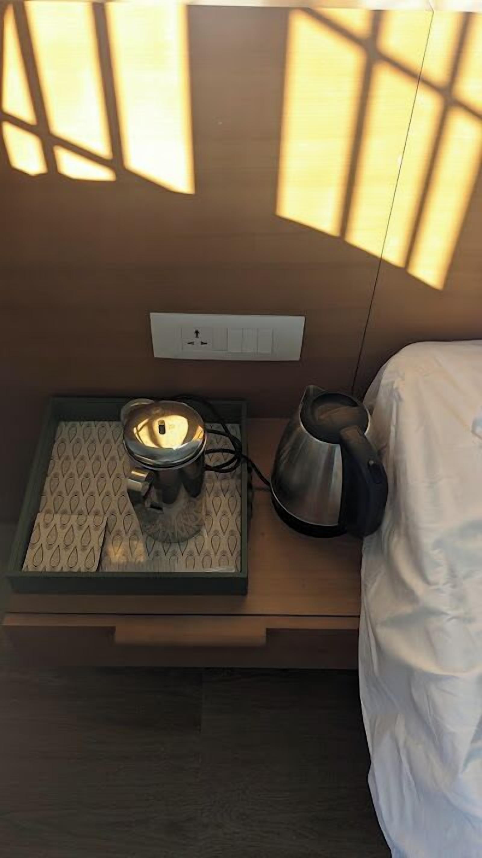 A neatly arranged hotel room with a bed, a bedside table with a kettle and a lamp, and a power outlet on the wall.