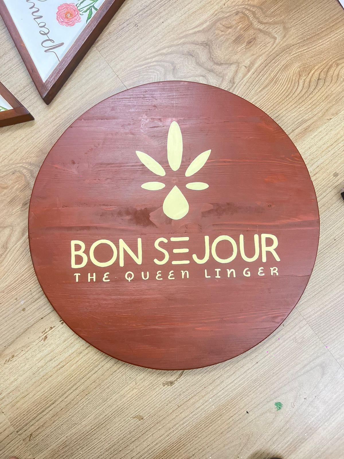 A circular wooden sign with the text "BON SEJOUR THE QUEEN LINGER" and a stylized flower design.