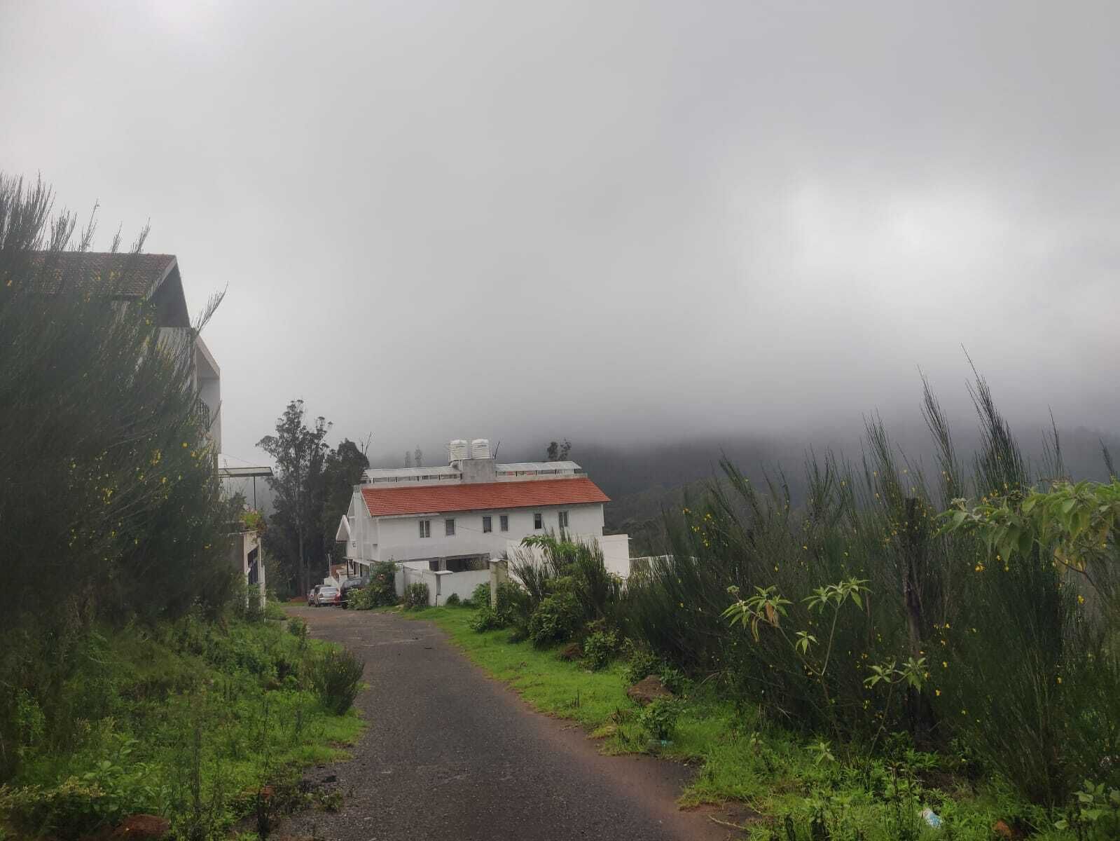 Eine Landstraße führt zu einem weißen Gebäude mit rotem Dach, umgeben von Grünflächen und nebelverhangenen Bergen im Hintergrund.