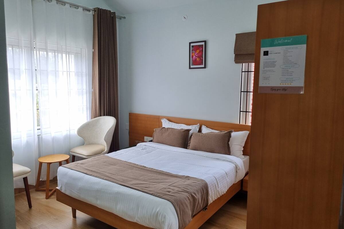 A hotel room with a wooden bed, a chair, a small table, and a window with curtains.