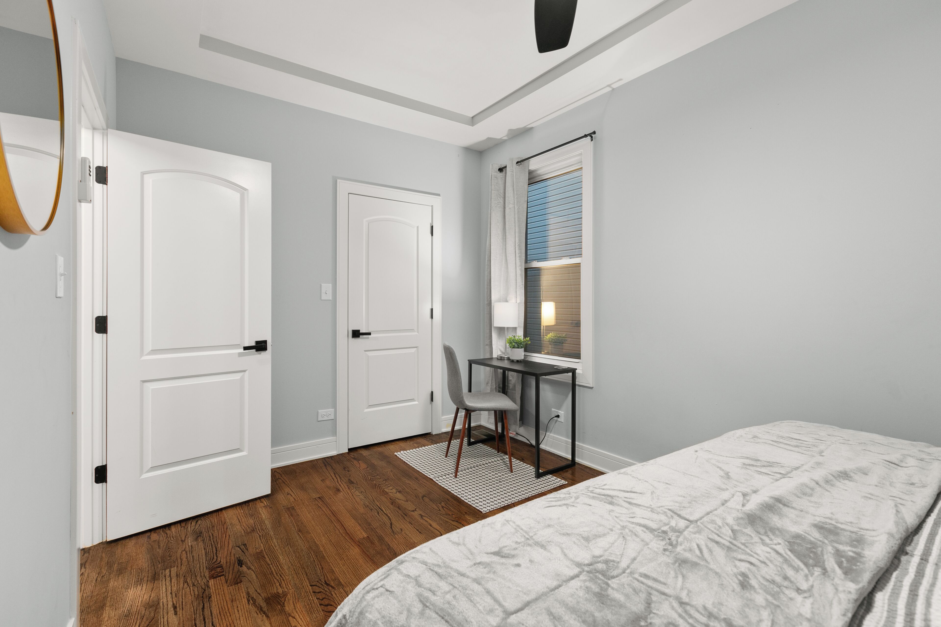 A bedroom with a bed, a desk with a chair, a window with blinds, and a door.