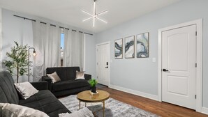 A modern living room with a black sofa, a round coffee table, and a plant. There are three framed artworks on the wall and a white door. - Luxury 5Bed 2Bath Apt w/Parking (Chicago)