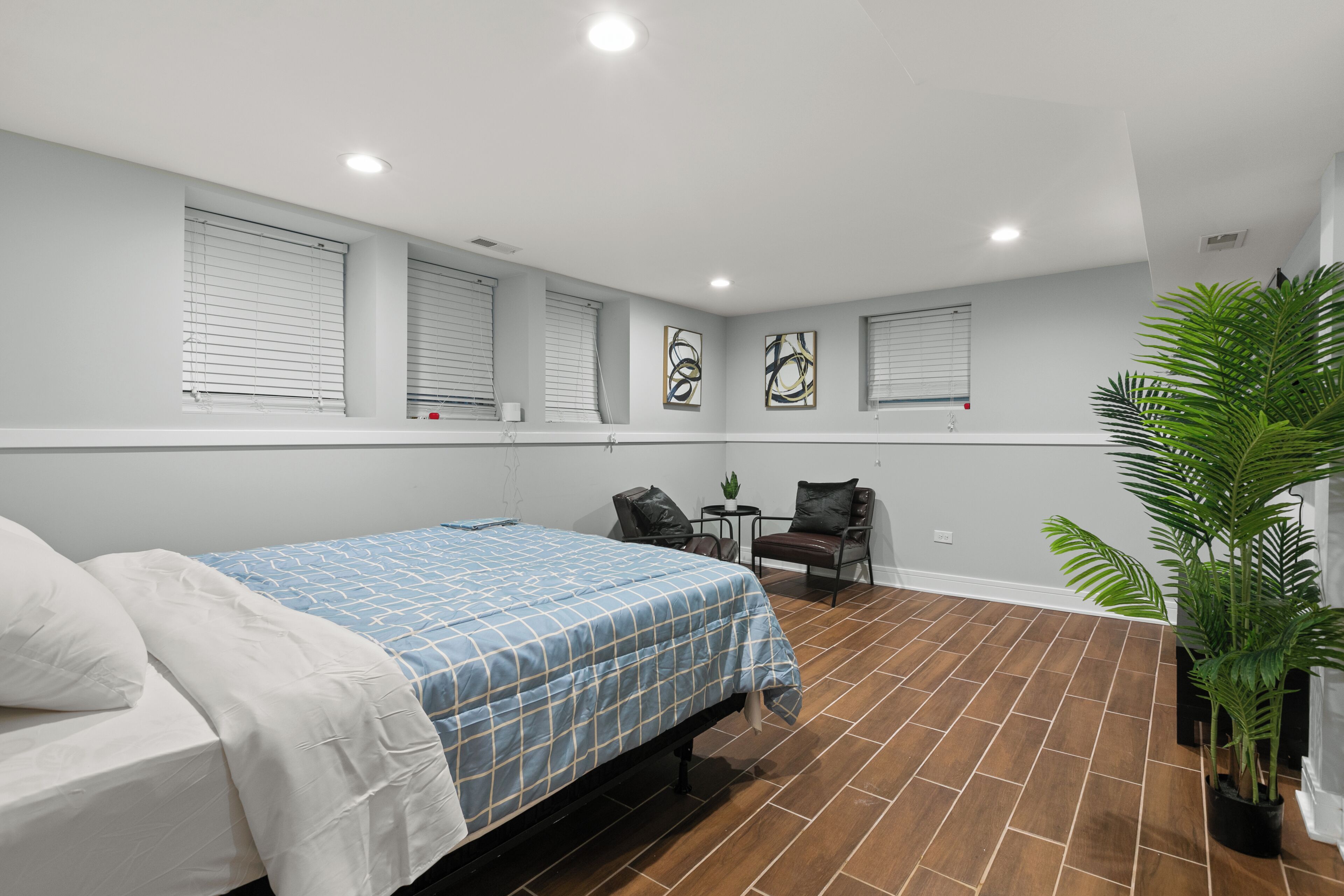 A bedroom with a bed, a small table, two chairs, a potted plant, and artwork on the walls.