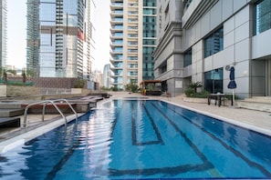 A swimming pool with clear blue water, surrounded by modern buildings and a covered seating area. - Impressive 2BR at Princess Tower (Dubai)