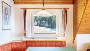 Una stanza con soffitto in legno, una finestra vista foresta, un tavolo con tovaglia verde e una panca rossa con motivo.