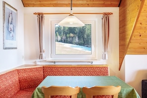 Une pièce avec un plafond en bois, une fenêtre donnant sur une forêt, une table recouverte d’une nappe verte et un banc rouge à motifs.