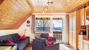 Una camera accogliente con soffitto in legno, un divano con cuscini rossi, una poltrona, un'area pranzo con pensili in legno e vista sugli alberi attraverso una finestra.