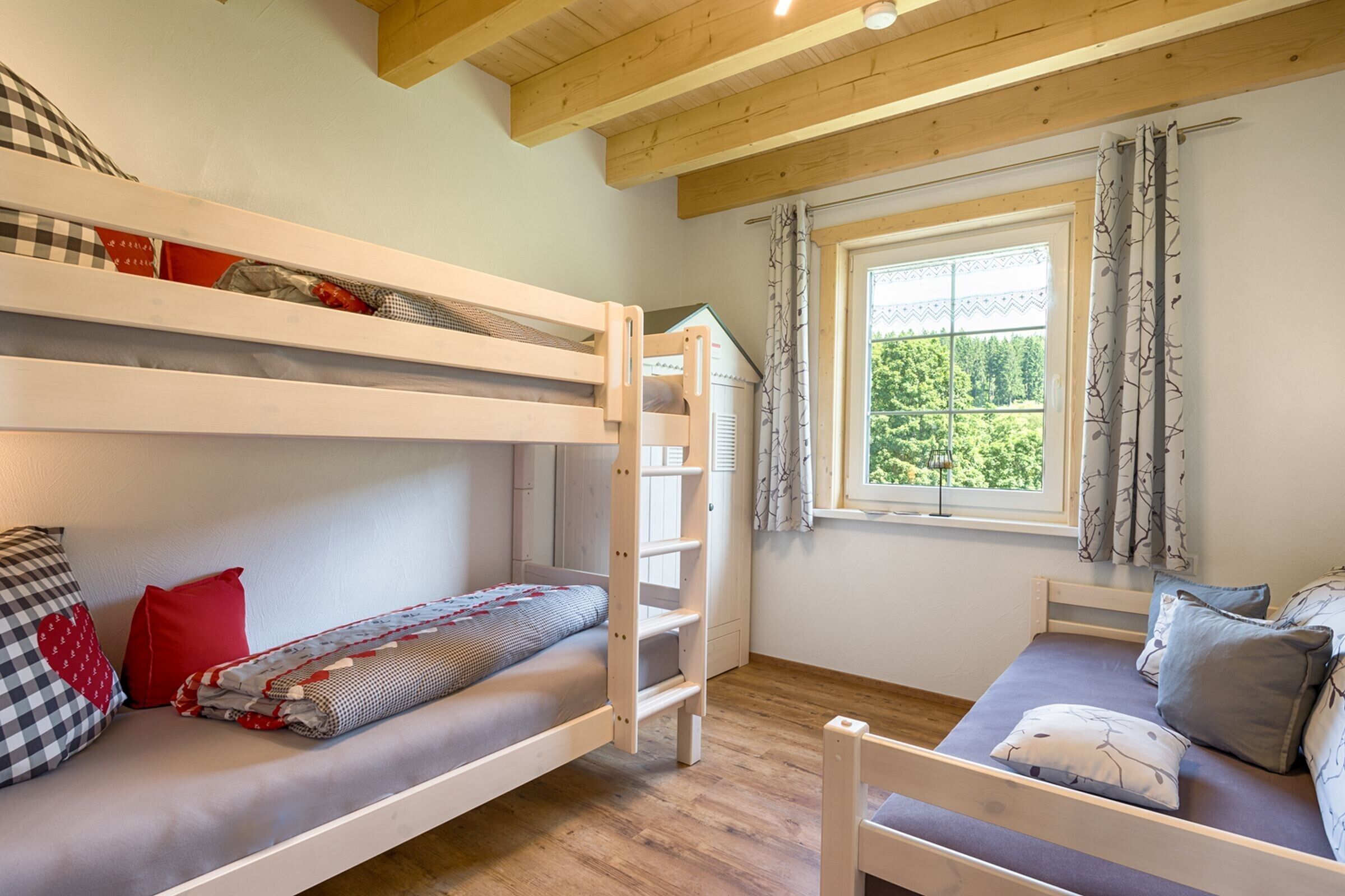 Ein Zimmer mit Etagenbetten, einer Schlafcouch, einem Fenster mit Vorhängen und Blick auf Grün.
