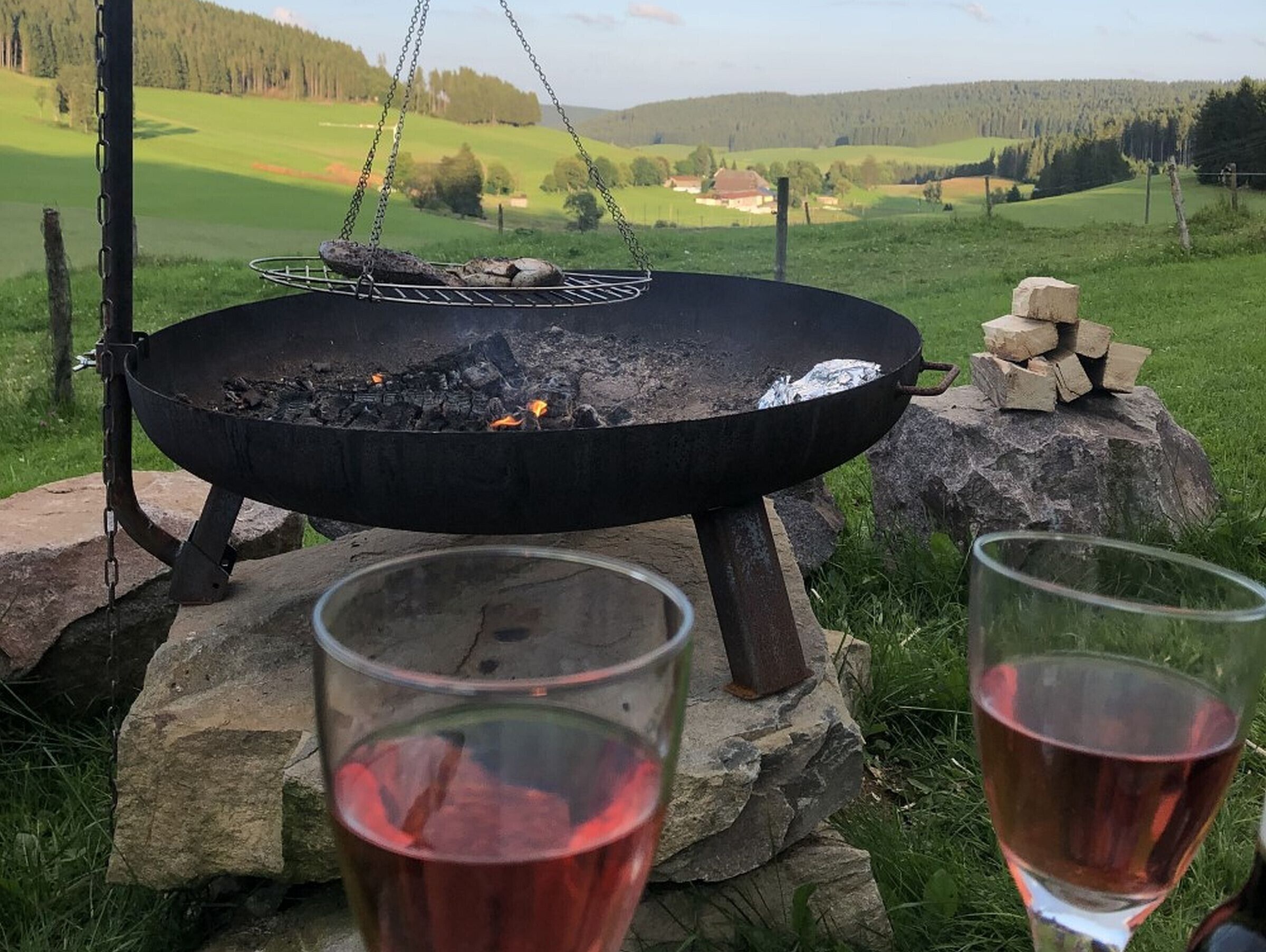 Ein Lagerfeuer mit Holzscheiten, einem hängenden Grill und zwei Gläsern Rotwein auf Steinen.