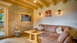 Una sala de estar acogedora con pared de piedra, vigas de madera, un sofá de cuero, una mesa de centro de madera con fruta, y un televisor de pantalla plana montado en la pared.