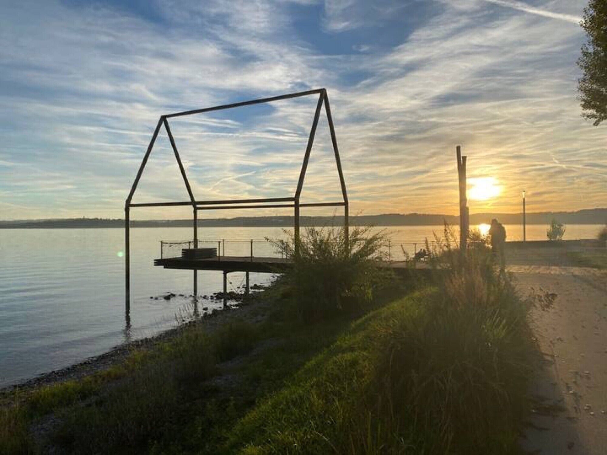 Blick auf ein Seeufer mit einer Metallkonstruktion, möglicherweise einem Pavillon, am Wasser.