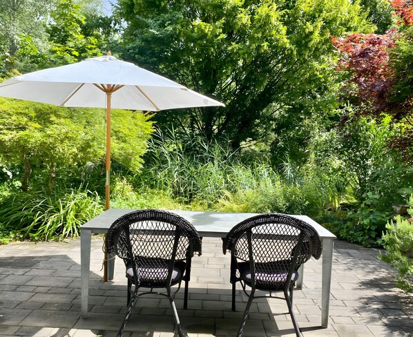 A garden patio with a table and two chairs, surrounded by lush greenery.