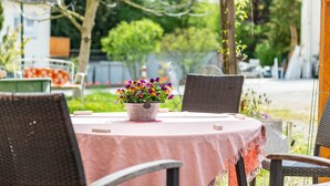 Un espacio de comedor al aire libre con sillas de mimbre y una mesa redonda cubierta con un mantel rosa. Hay una planta en maceta con flores moradas y anaranjadas sobre la mesa.