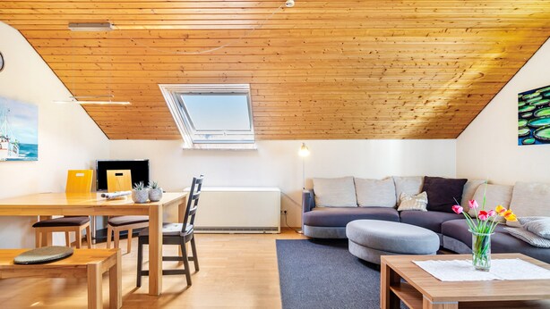 Un loft moderno con techo inclinado, una zona de comedor con muebles de madera, un conjunto de sofás y un cuadro en la pared.