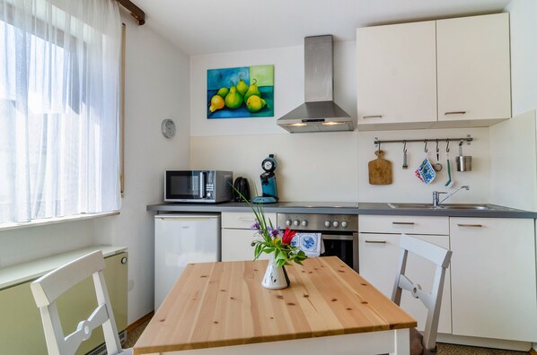 Una cocina compacta con armarios blancos, un microondas, un hervidor y una estufa. Hay una mesa de madera con un jarrón de flores.
