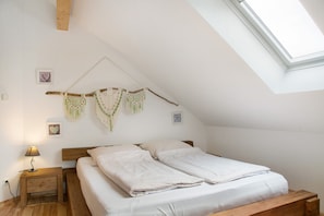 Quarto com teto inclinado, cama com roupa de cama branca, mesa de cabeceira de madeira com abajur e um pendente de macramé na parede.
