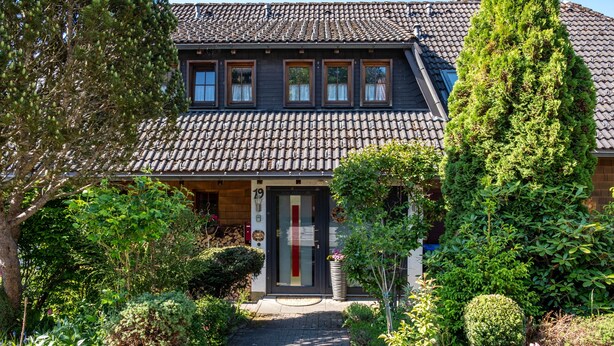Ett tvåvåningshus med ett tak täckt av tegelpannor, en ytterdörr med en röd rand och en trädgård med olika växter och ett högt träd.