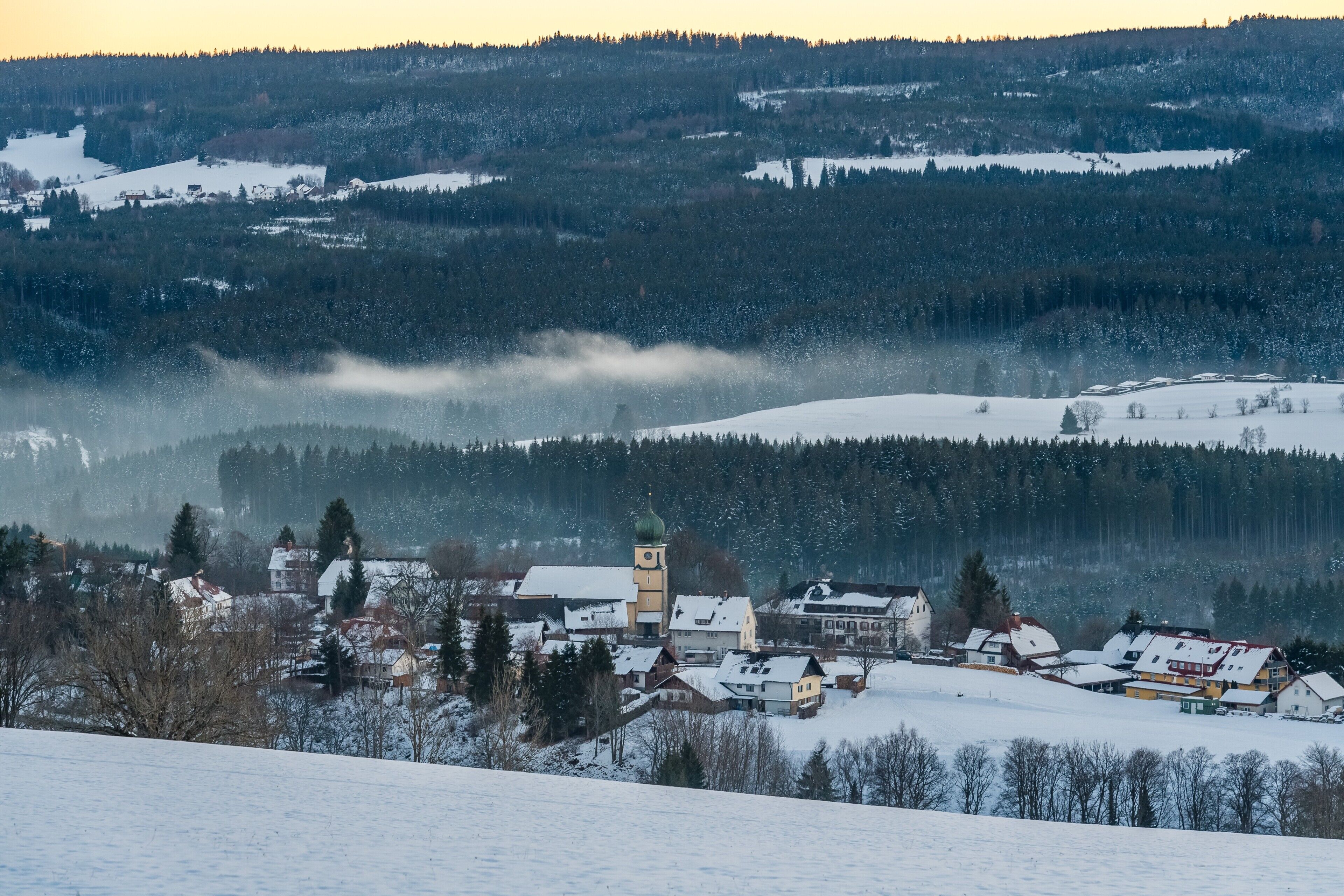 A snow-covered village with a prominent church tower, surrounded by forested hills.