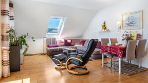 Ett modernt vardagsrum med en soffa, en svart läderfåtölj, ett matbord med stolar, en krukväxt och ett fönster med utsikt.