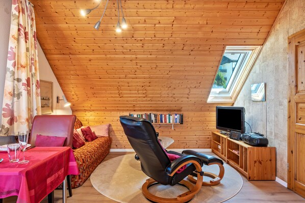 A cozy room with a wooden ceiling, a rocking chair, a television, and a bookshelf. - Apartment 'Feldberg' with Mountain View, Southwest Balcony, Table Tennis, and Wi-Fi (Lenzkirch)