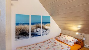 Quarto com teto inclinado, cama com roupa de cama floral, luminária fixada na parede e uma obra de arte com tema praiano.