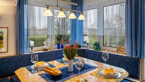 Uma área de jantar com uma mesa posta para dois, um vaso de flores e vista para o exterior.