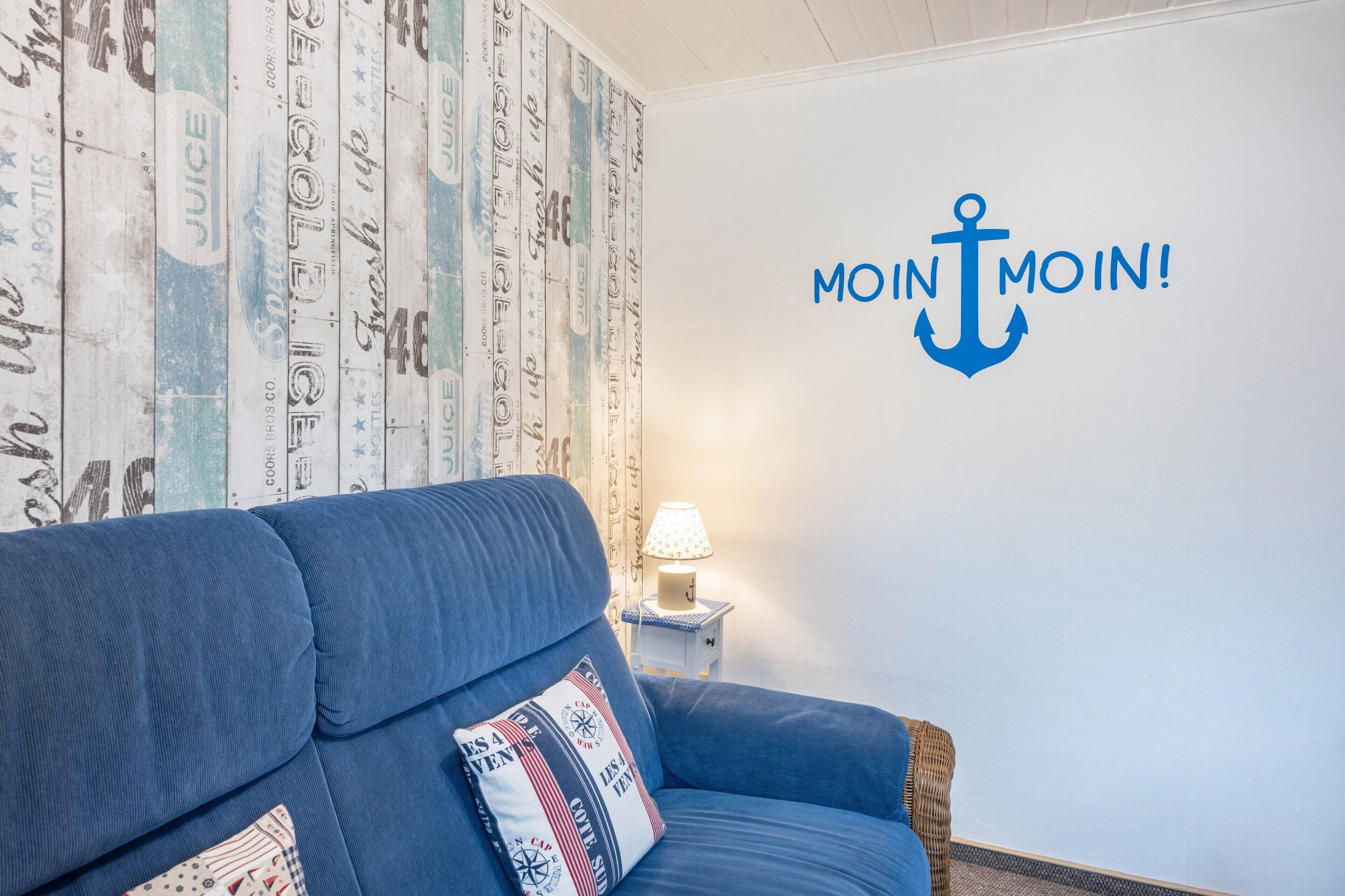 Un canapé bleu orné d’un coussin sur le thème de la marine, un mur décoré d’une grande ancre et de l’inscription « MOIN MOIN ! », et une petite table avec une lampe.