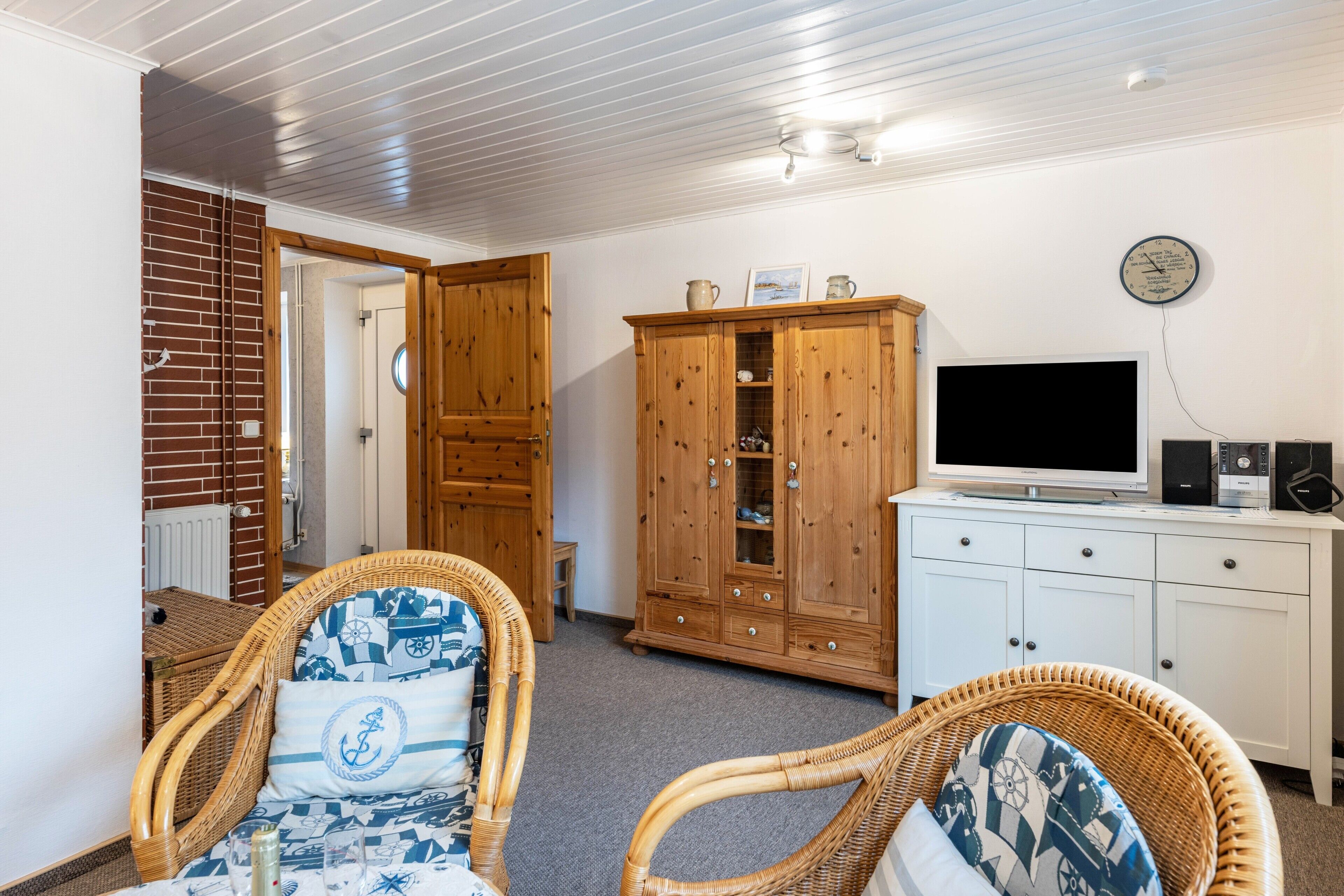 Ein Zimmer mit Fernseher, Holzschränken, Korbstühlen und einer Wanduhr.