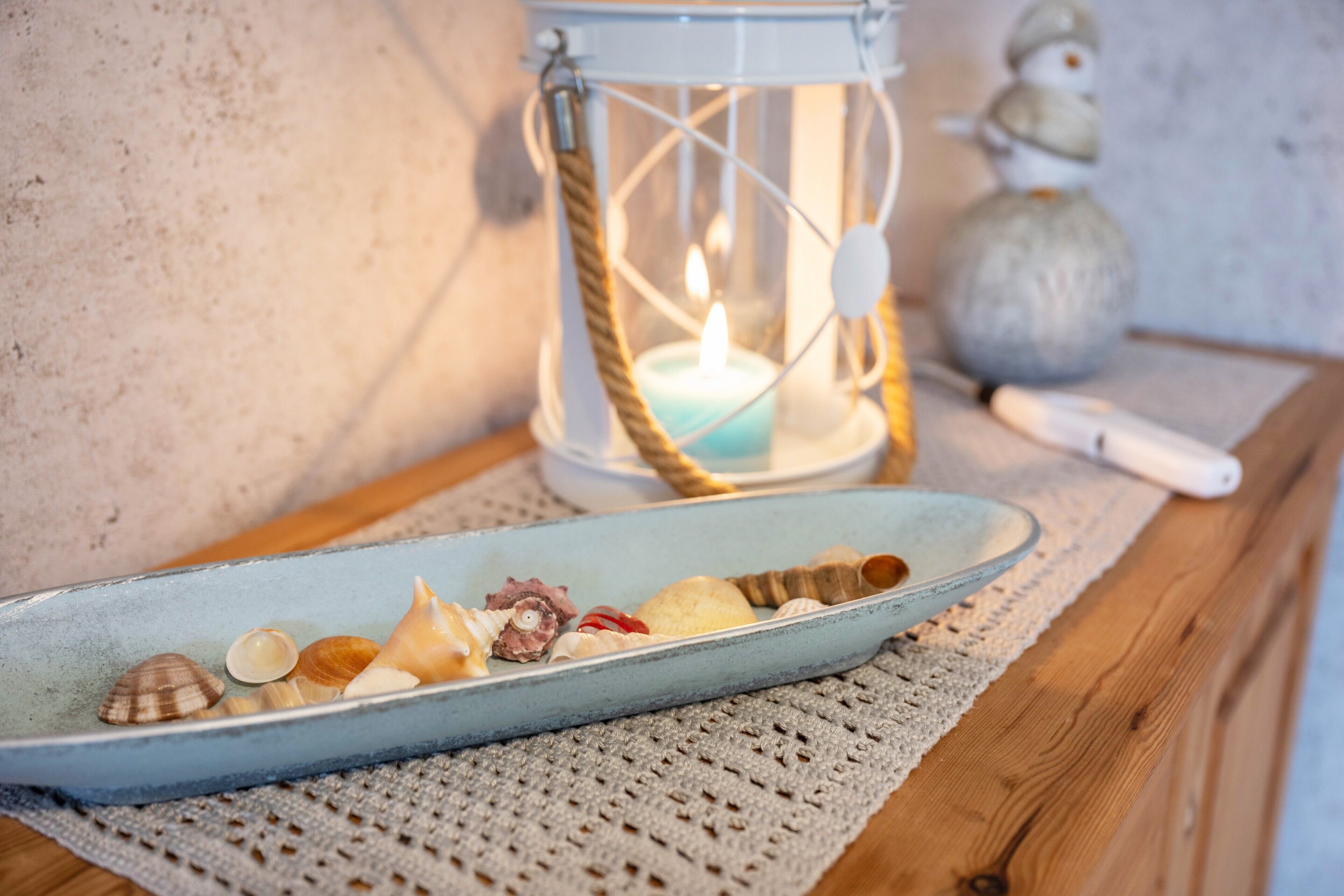 A candle in a lantern, a snowman figurine, and a ceramic plate with seashells and a conch shell on a wooden table.