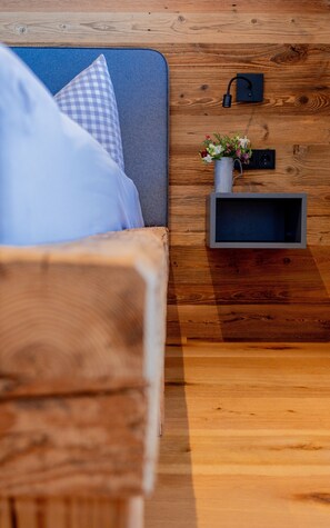 A wooden floor, a bed with a grey headboard, a small shelf with a vase of flowers, and a wall-mounted speaker. - Apartment 'Wiagsog' with Mountain View, Balcony and Wi-Fi (Garmisch-Partenkirchen)
