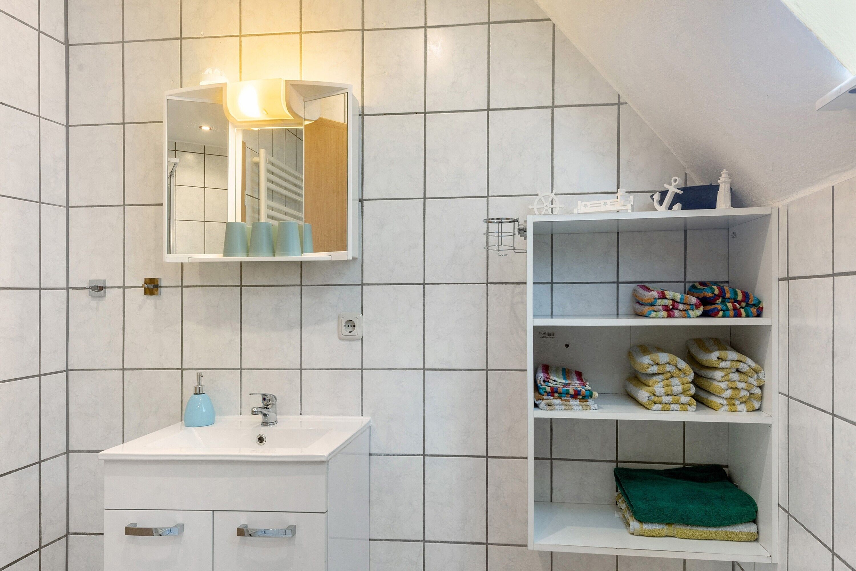 Et badeværelse med hvid vask, spejl og hylder med håndklæder og toiletartikler.