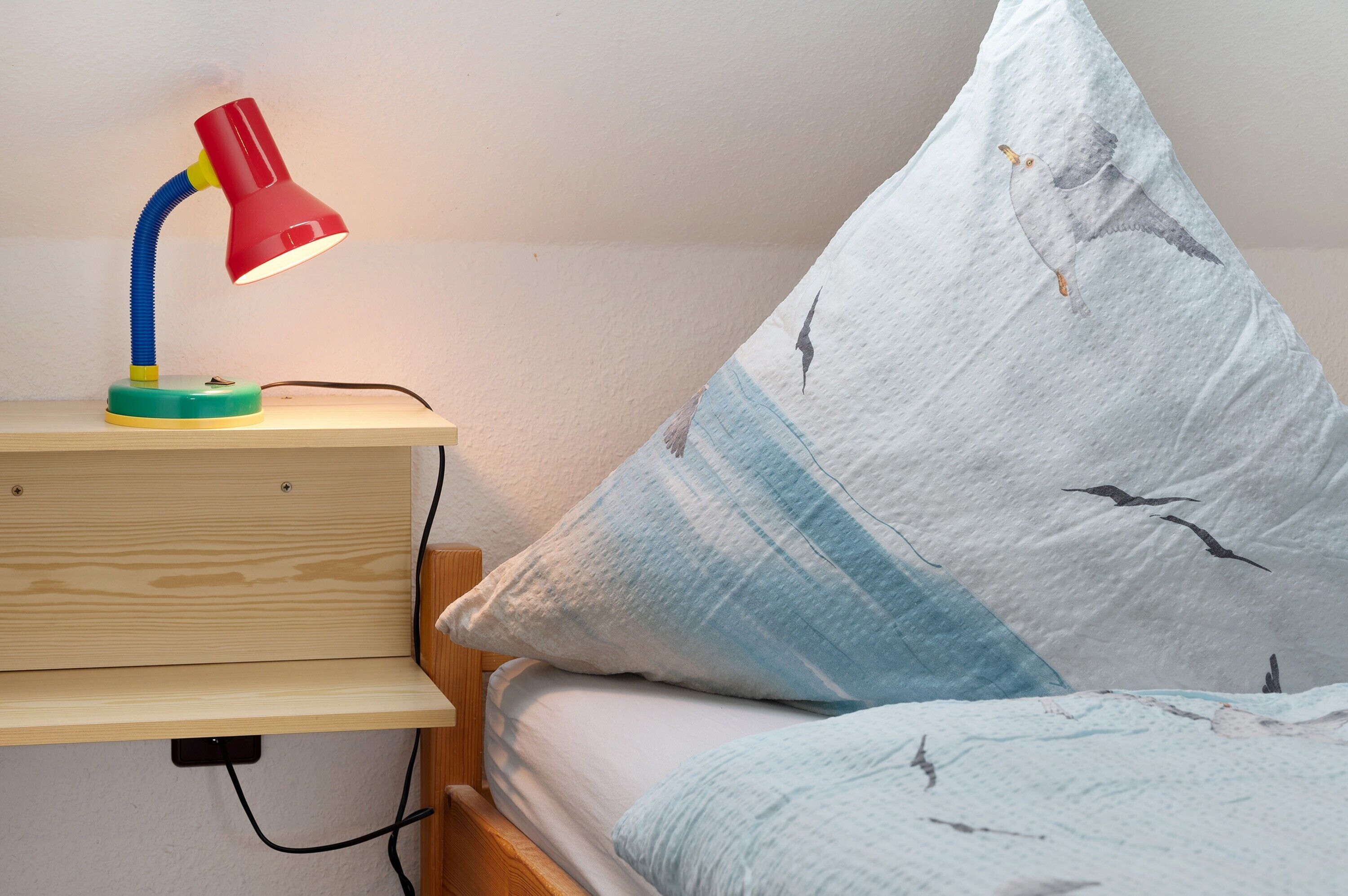 Een nachtkastje met een rode bureaulamp, een kussen met een vogelprint, en een houten nachtkastje.