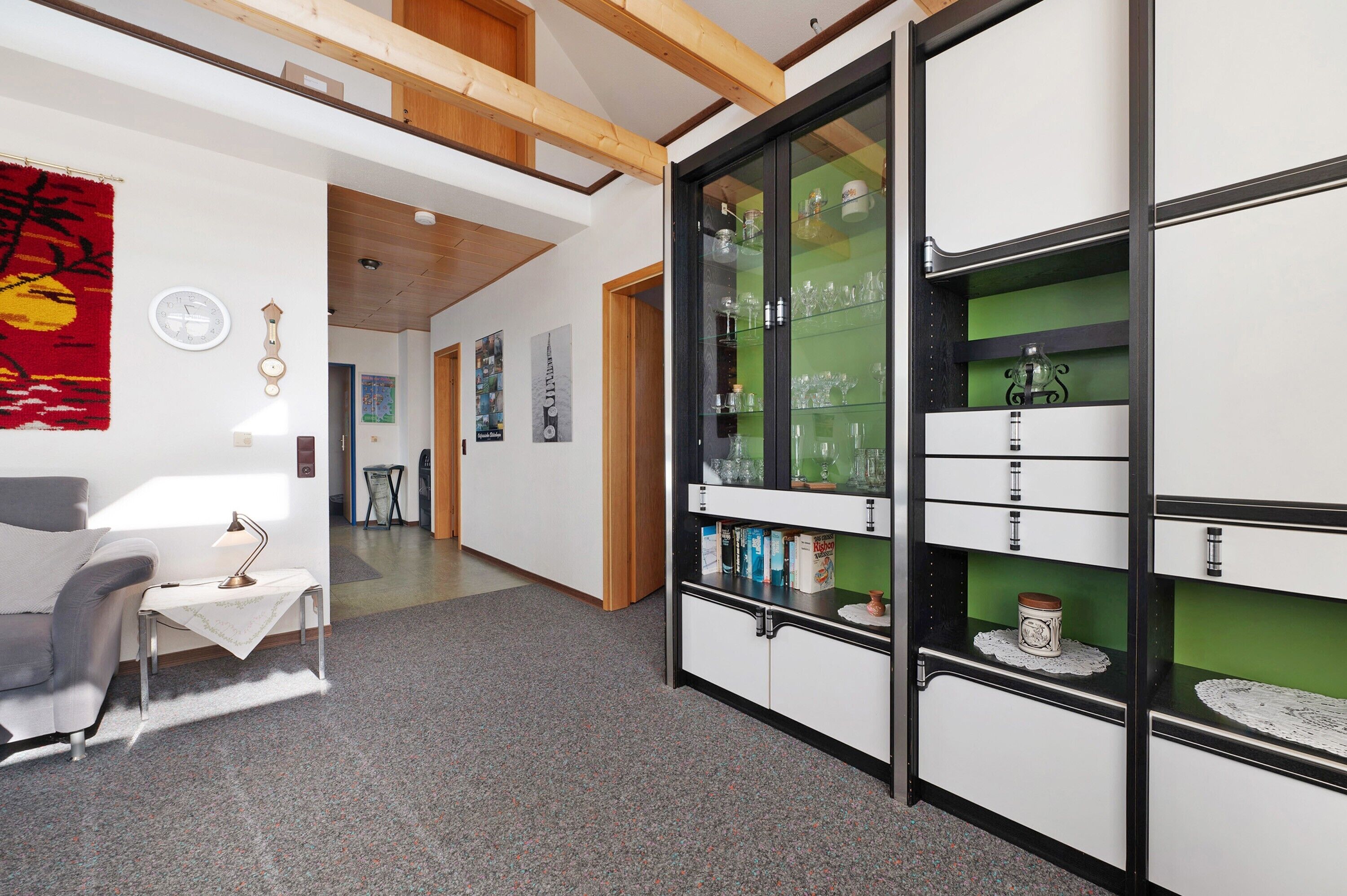 Een kamer met een bank, een klein tafeltje met een lamp, een kast met glazen deuren en een rode wandtapijt.
