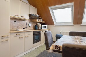 A compact kitchen with a stove, microwave, and sink, a dining table with chairs, and a window with blinds.