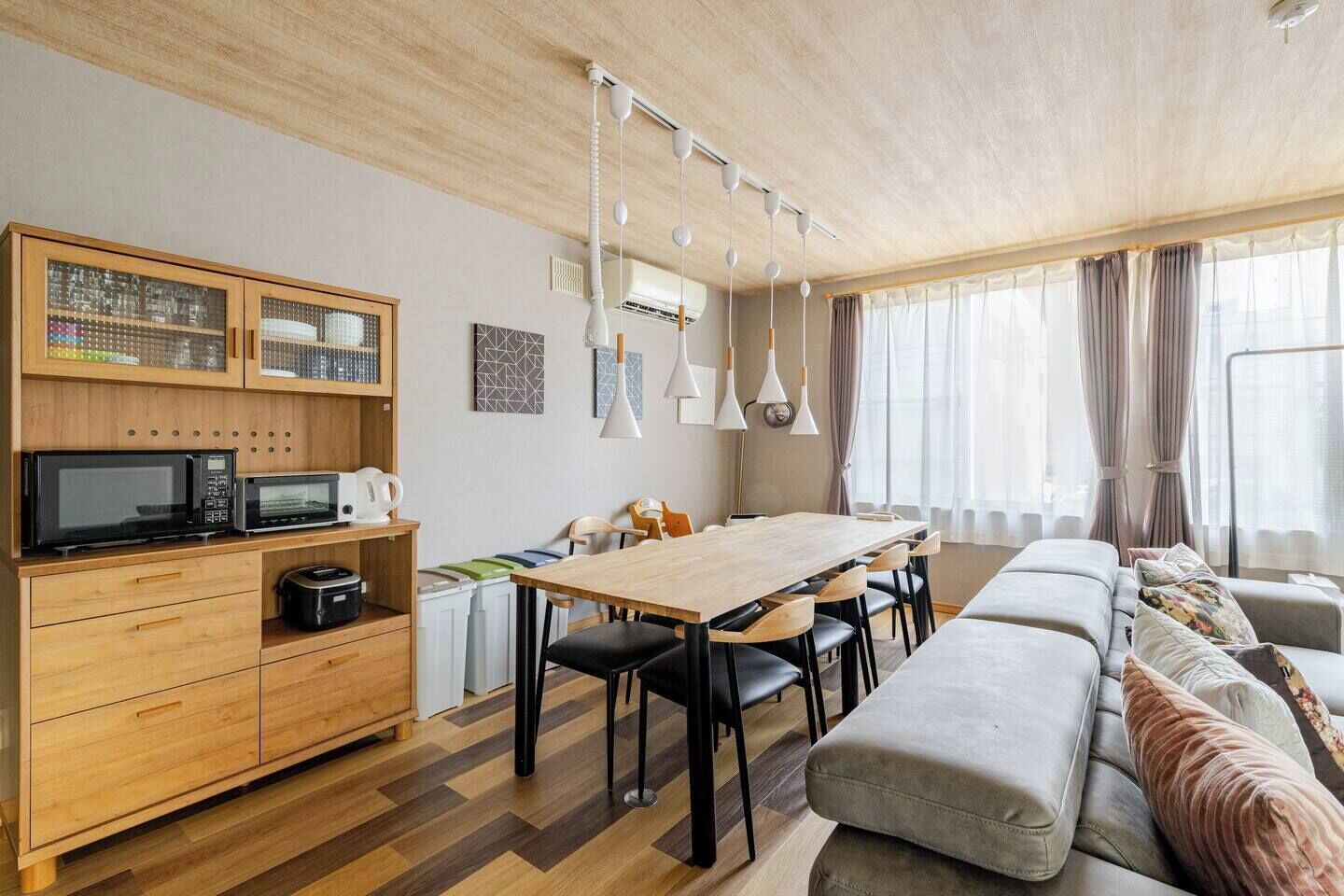 A modern living room with a wooden dining table, black chairs, a sofa, and a kitchen area with a microwave and cabinets.