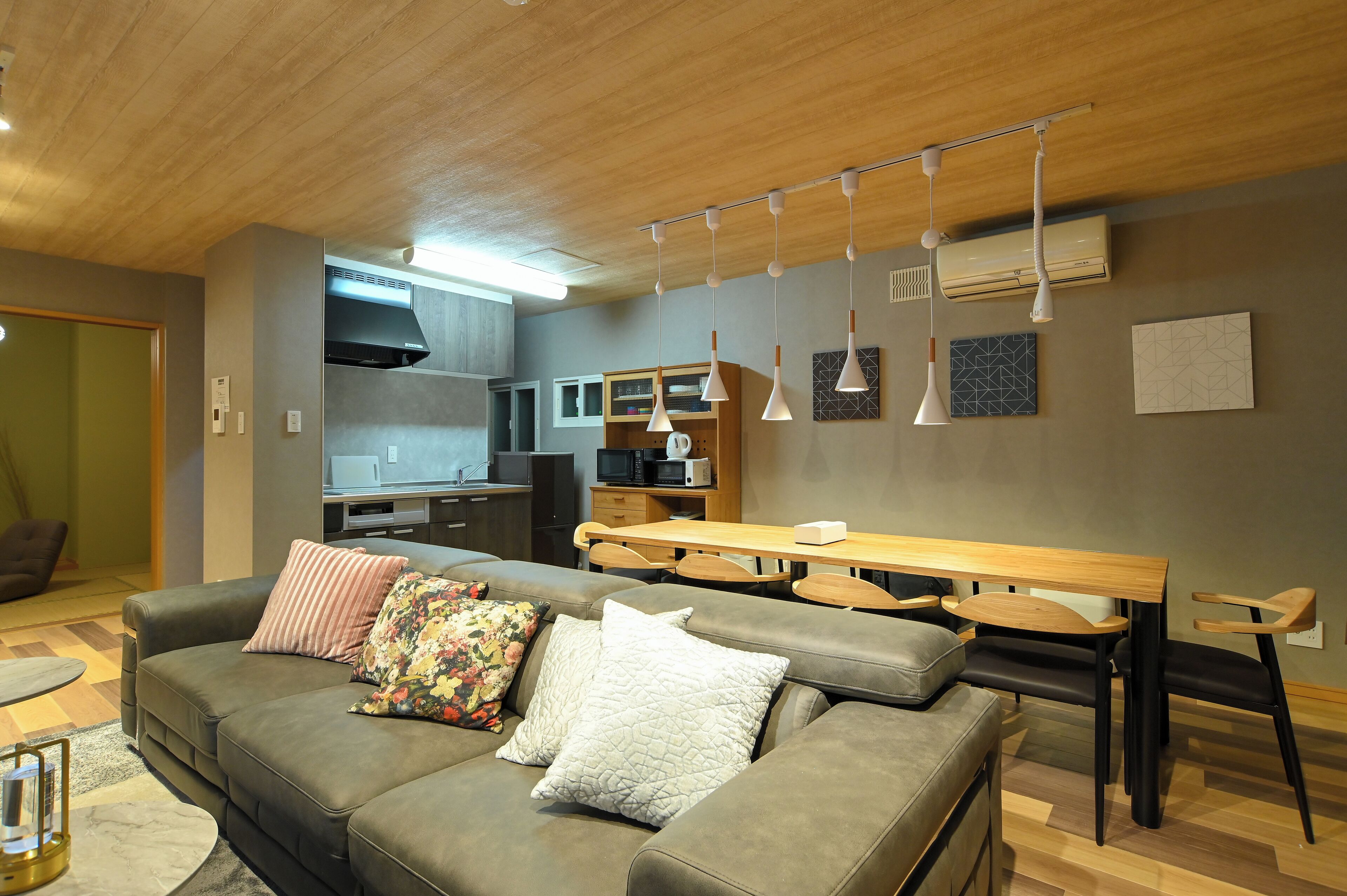 A modern living room with a grey sofa, a wooden dining table, and a kitchen in the background.