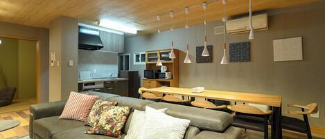 A modern living room with a grey sofa, a wooden dining table, and a kitchen in the background.
